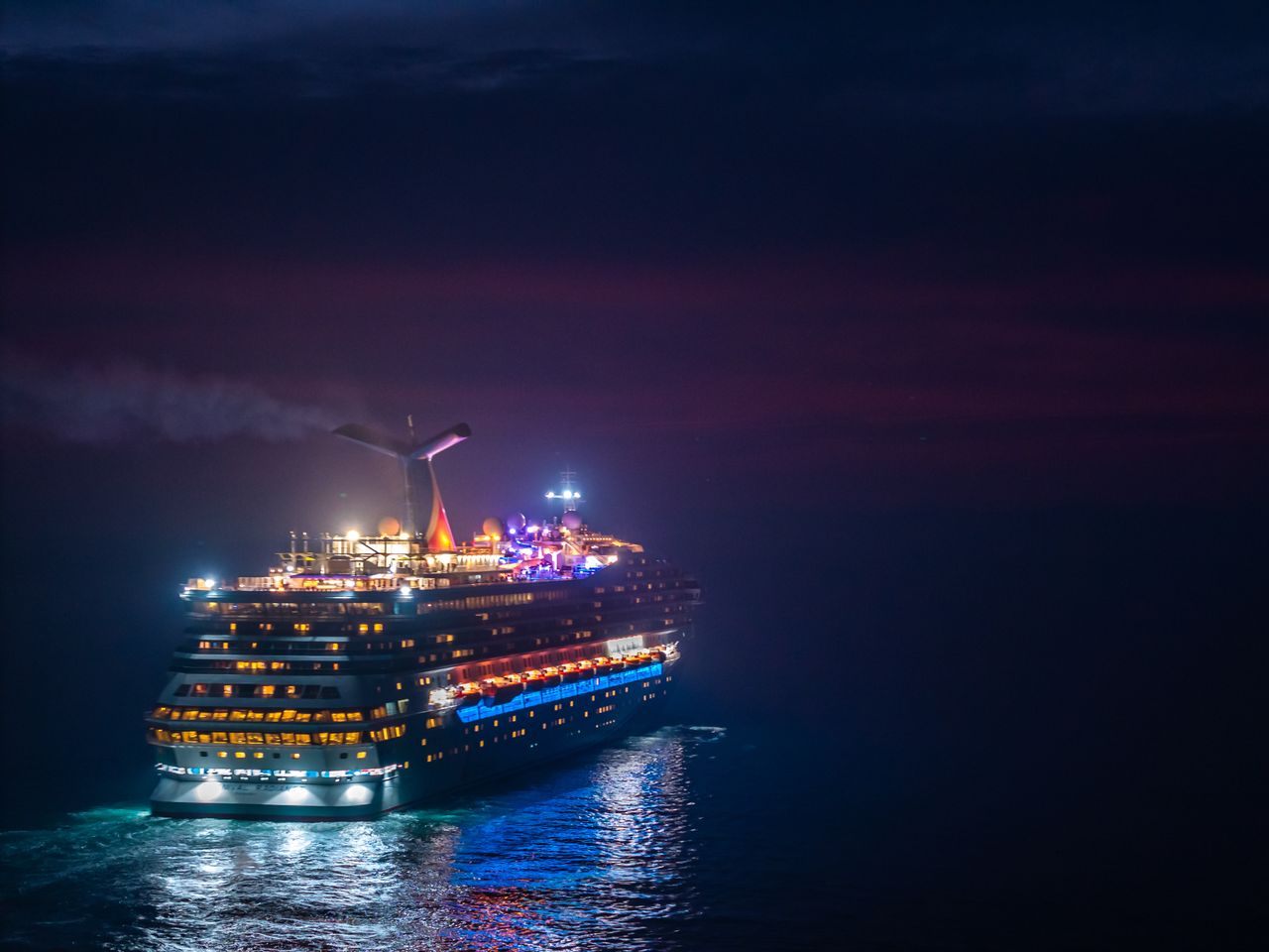 services-02 A brightly lit cruise ship sails through the ocean under a vivid night sky.