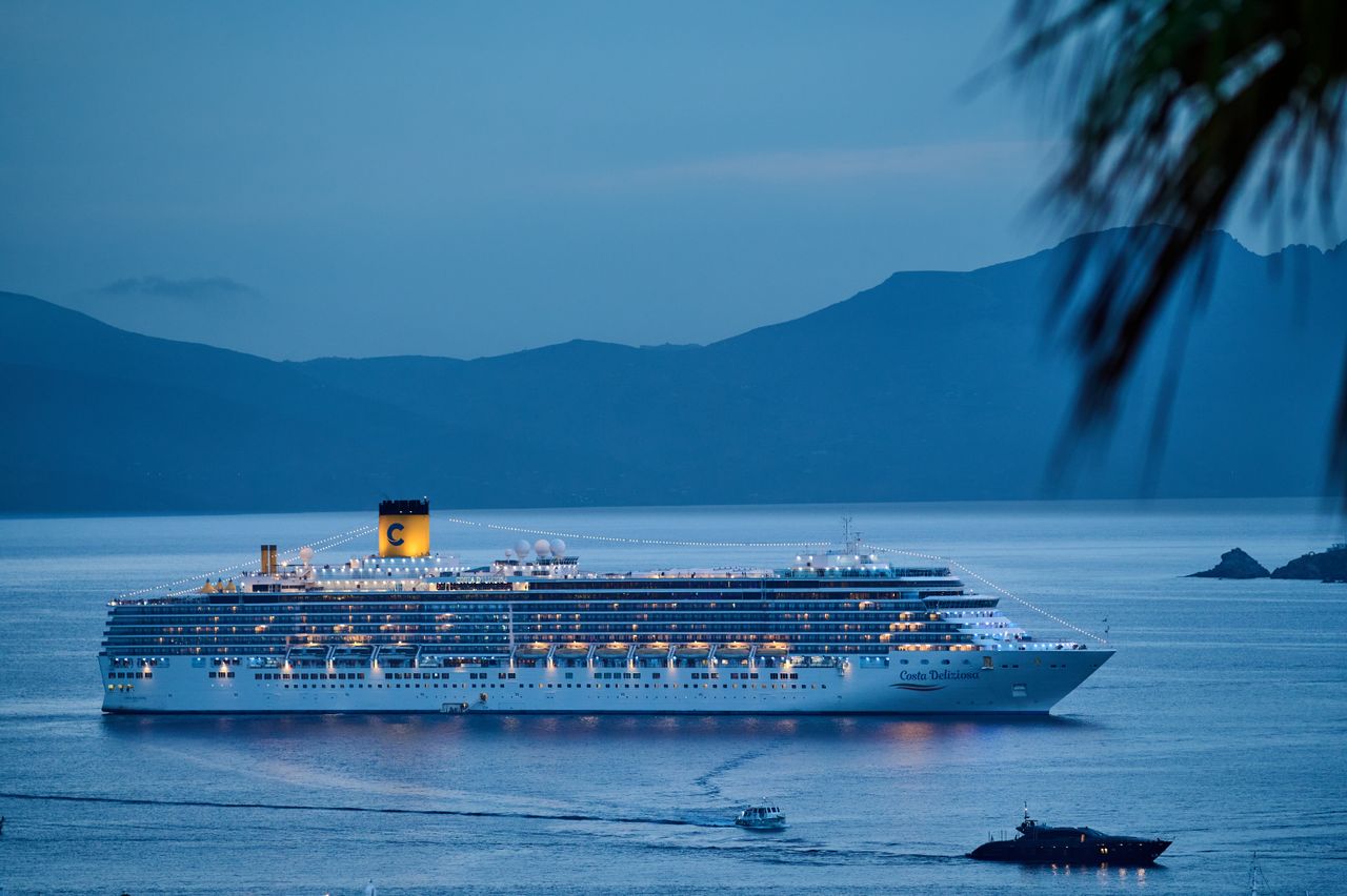 services-03 A beautifully illuminated cruise ship sails across the calm sea at dusk, framed by mountains.